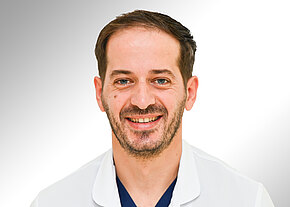 Porträt eines lächelnden Arztes in weißem Kittel vor neutralem Hintergrund.