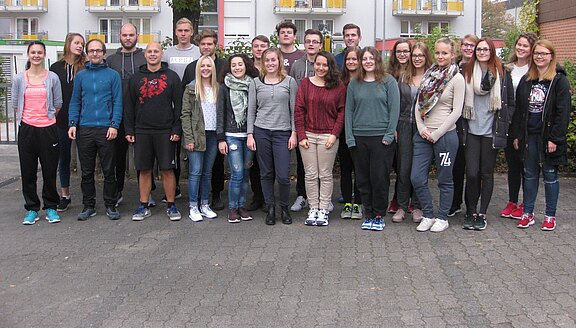 Eine größere Gruppe junger Erwachsener steht dicht beieinander und posiert lächelnd für ein Gruppenfoto vor einem Gebäude mit Balkonen, im Freien, in Freizeitkleidung.