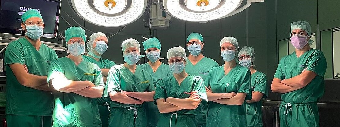Eine Gruppe von zehn Fachleuten im Operationssaal steht in grüner Arbeitskleidung und Masken. Sie posieren entschlossen vor einem chirurgischen Tisch, umgeben von medizinischen Geräten und OP-Lichtern. Die Atmosphäre wirkt professionell und konzentriert.