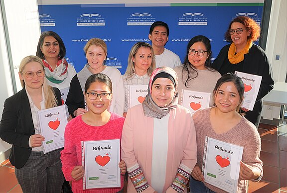 Gruppenfoto: Zehn Personen lächeln und halten Urkunden mit großem rotem Herz und kleinem Kreuz. Sie feiern eine abgeschlossene Schulung oder Auszeichnung im Pflege‑/Gesundheitsbereich. Helle Innenraumszene, formelle Kleidung.
