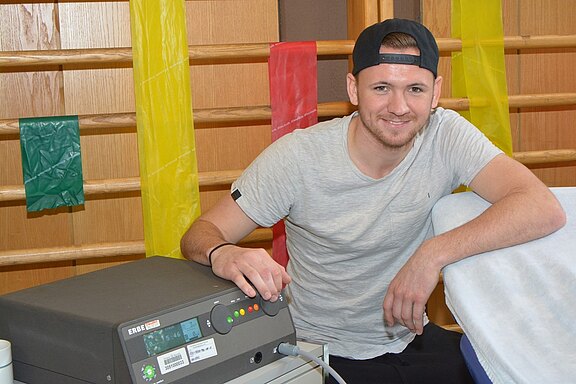 Ein junger Mann mit kurzer Frisur und einem Baseballcap sitzt an einem medizinischen Gerät und lächelt in die Kamera. Der Hintergrund ist mit farbigen Bändern geschmückt. Neben dem Gerät befinden sich ein Handgriff und ein Tisch mit einer blauen Decke.