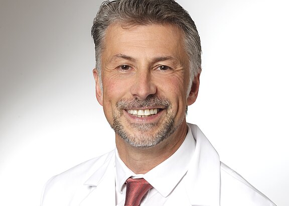 Ein lächelnder Mann trägt einen weißen Arztkittel und eine rote Krawatte. Sein Gesicht zeigt ein freundliches Auftreten. Der Hintergrund ist neutral gehalten, um den Fokus auf den Mann zu legen.