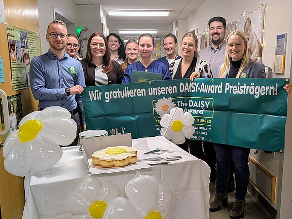 Gruppe von Klinikmitarbeitenden in einem Flur hält ein grünes Banner: „Wir gratulieren unseren DAISY‑Award Preisträgern!“. Davor ein Tisch mit Torte, Tellern und weißen Gänseblümchen-Luftballons. Feierliche Stimmung; Auszeichnung für Pflegekräfte wird gewürdigt.