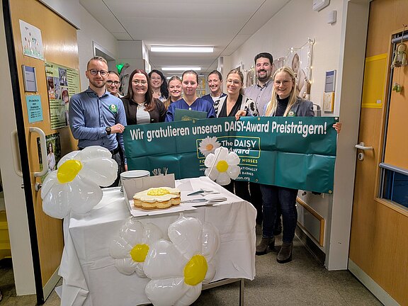 Gruppe von Klinikmitarbeitenden in einem Flur hält ein grünes Banner: „Wir gratulieren unseren DAISY‑Award Preisträgern!“. Davor ein Tisch mit Torte, Tellern und weißen Gänseblümchen-Luftballons. Feierliche Stimmung; Auszeichnung für Pflegekräfte wird gewürdigt.