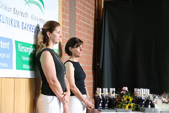 Zwei Frauen in schwarzen Oberteilen und weißen Hosen stehen neben einem Tisch mit verpackten Flaschen, Gläsern und einem Blumenstrauß. Im Hintergrund ein Schild mit der Aufschrift „Klinikum Bayreuth“. Die Szene wirkt wie eine Empfangs- oder Veranstaltungs­situation.