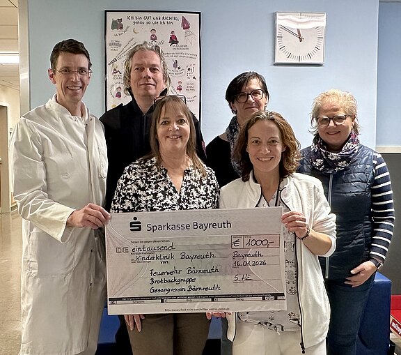 Sechs Personen in einem Krankenhausflur, zwei im Arztkittel, halten gemeinsam einen übergroßen Spendenscheck der Sparkasse Bayreuth. Der Scheck über 1.000 Euro ist für das Klinikum Bayreuth bestimmt. Die Gruppe lächelt in die Kamera.