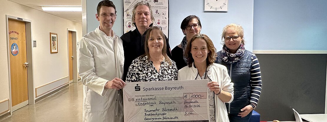 Sechs Personen in einem Krankenhausflur, zwei im Arztkittel, halten gemeinsam einen übergroßen Spendenscheck der Sparkasse Bayreuth. Der Scheck über 1.000 Euro ist für das Klinikum Bayreuth bestimmt. Die Gruppe lächelt in die Kamera.
