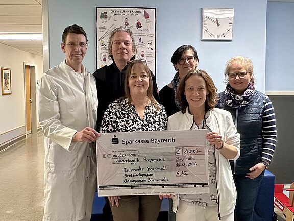 Sechs Personen in einem Krankenhausflur, zwei im Arztkittel, halten gemeinsam einen übergroßen Spendenscheck der Sparkasse Bayreuth. Der Scheck über 1.000 Euro ist für das Klinikum Bayreuth bestimmt. Die Gruppe lächelt in die Kamera.