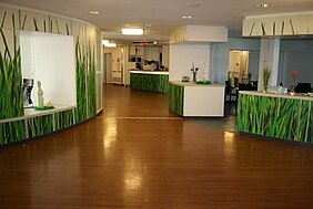 Der Raum zeigt einen hellen Empfangsbereich mit abgerundeten Wänden, die mit grünem Grasdesign dekoriert sind. Der Boden ist aus Holz und die Einrichtung ist modern. Im Hintergrund sind einige Räume und eine Empfangstheke sichtbar.