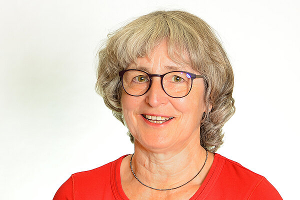 Porträt einer lächelnden Frau mit grauen Haaren und Brille, sitzend, in rotem Oberteil vor hellem neutralem Hintergrund.