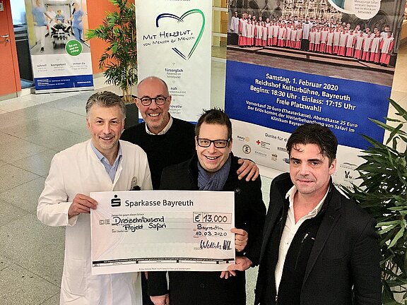 Vier Männer stehen zusammen und halten einen großen Scheck der Sparkasse Bayreuth über 13.000 Euro, der für das Projekt "Safari" bestimmt ist. Im Hintergrund sind zwei Banner zu sehen, die für ein Benefizkonzert der Regensburger Domspatzen werben.