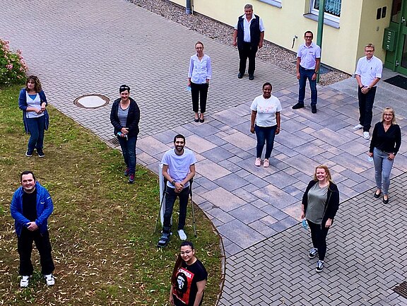 Gruppe von etwa zwölf Erwachsenen steht im Freien auf einem Hof, mit Abstand zueinander, blickt zur Kamera und lächelt. Aus erhöhter Perspektive aufgenommen. Sie bilden ein lockeres Raster auf Pflasterfläche und Rasen. Stimmung freundlich, eine Art Team- oder Kursfoto.