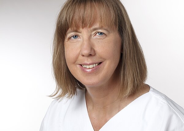 Porträt einer lächelnden Frau mit schulterlangem hellbraunem Haar und Pony, in weißer Kleidung vor neutralem, hellem Hintergrund.