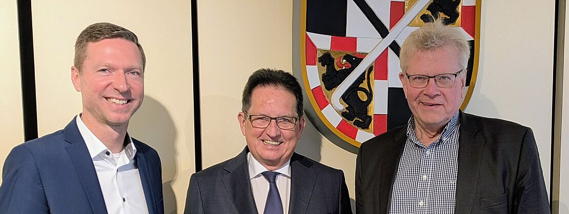 Drei Männer stehen in Anzug und Hemd nebeneinander vor einem Wappen. Sie lächeln freundlich in die Kamera. Der Hintergrund ist neutral gehalten. Die Szene vermittelt ein Gefühl von Zusammenarbeit oder offiziellem Anlass.