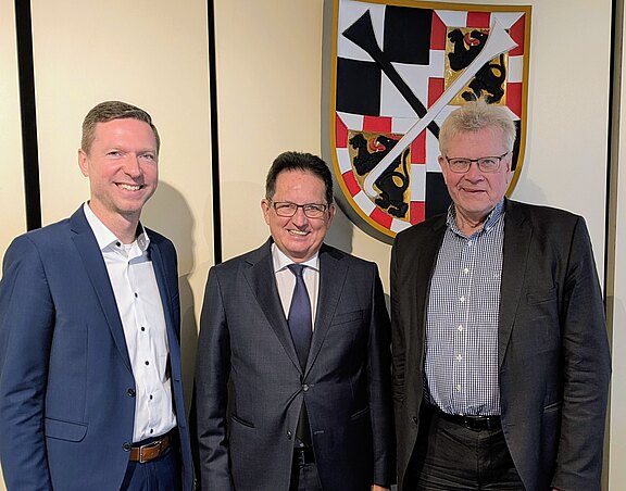 Drei Männer stehen in Anzug und Hemd nebeneinander vor einem Wappen. Sie lächeln freundlich in die Kamera. Der Hintergrund ist neutral gehalten. Die Szene vermittelt ein Gefühl von Zusammenarbeit oder offiziellem Anlass.