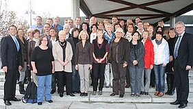 Eine Gruppe von etwa 50 Personen steht auf einer Treppe unter einem Vordach. Die Anwesenden lachen und posieren für ein Foto. Sie sind vielfältig gekleidet, einige tragen Jacken, andere Blusen oder Anzüge. Die Stimmung wirkt freundlich und gemeinschaftlich.