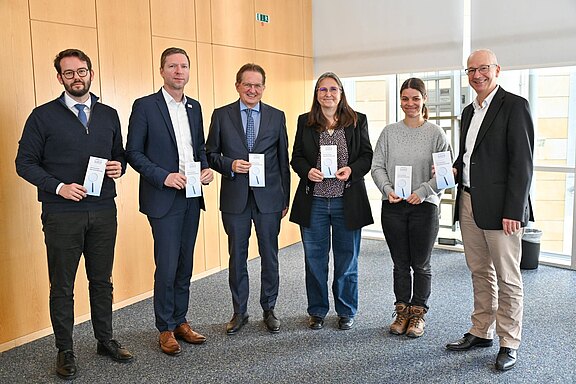 Sechs Erwachsene posieren als Gruppe in einem hellen Konferenzraum, alle halten Informationsflyer und lächeln in die Kamera; Mischung aus Anzug- und Freizeitkleidung, offizieller Anlass.