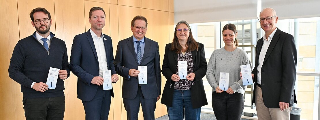 Sechs Erwachsene posieren als Gruppe in einem hellen Konferenzraum, alle halten Informationsflyer und lächeln in die Kamera; Mischung aus Anzug- und Freizeitkleidung, offizieller Anlass.