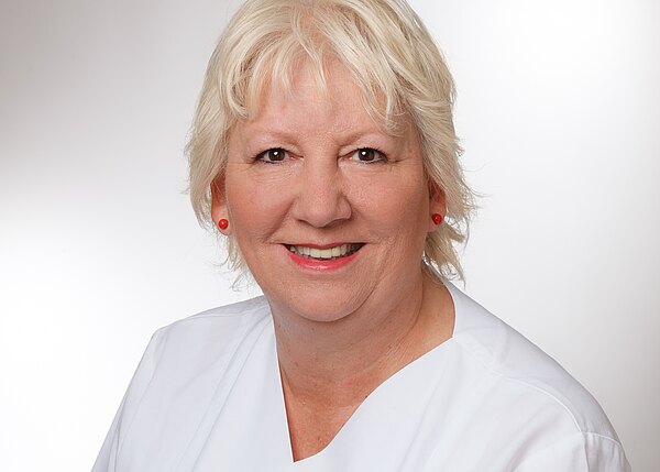 Porträt einer lächelnden älteren Frau mit kurzem blondem Haar, roten Ohrringen und Lippenstift, in weißer medizinischer Arbeitskleidung vor neutralem Hintergrund.