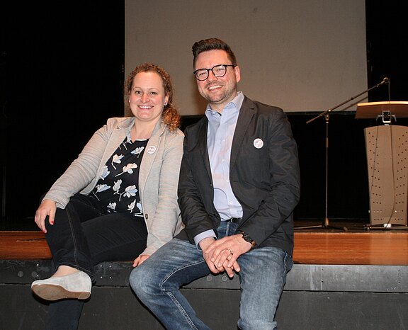 Zwei lächelnde Erwachsene sitzen entspannt am Bühnenrand. Die Person links trägt eine helle Strickjacke und Sneakers; die Person rechts trägt Brille, Blazer und Jeans. Im Hintergrund stehen ein Rednerpult und ein Mikrofon, was auf eine Veranstaltung oder Vortrag hinweist.