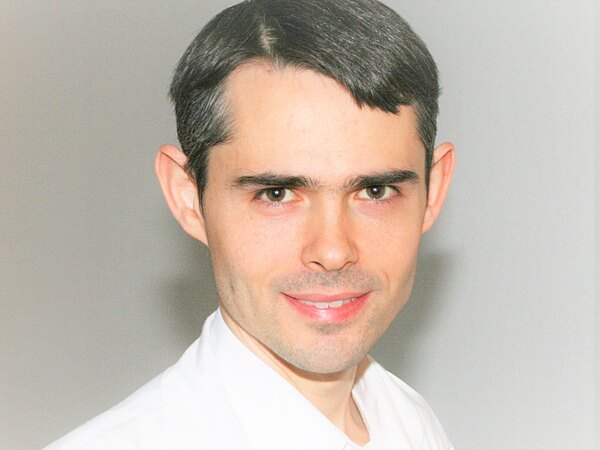 Porträtfoto: Nahaufnahme einer lächelnden Person mit kurzen dunklen Haaren, in weißem Hemd vor neutralem Hintergrund.