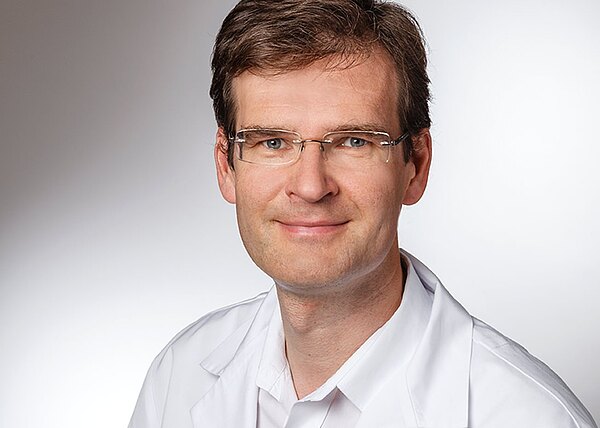 Porträtfoto: Ein freundlich lächelnder Mann mit kurzer brauner Frisur und Brille trägt einen weißen Kittel und blickt in die Kamera vor neutralem Hintergrund.