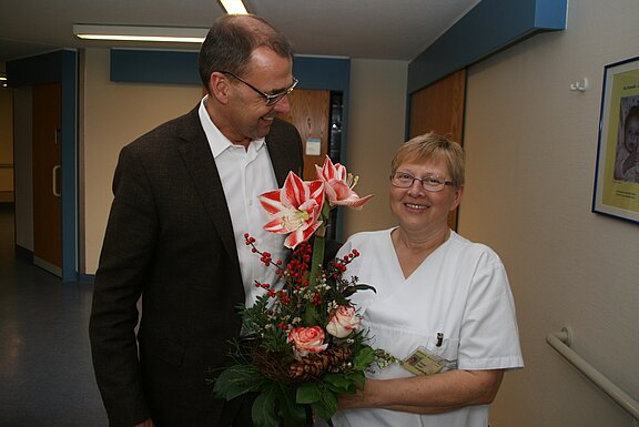 Eine lächelnde Pflegekraft hält einen großen Blumenstrauß; ein Mann im Anzug steht daneben und schaut sie freundlich an, im Flur eines Krankenhauses.