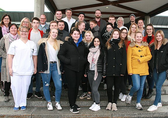 Gruppenfoto mit etwa 25 jungen Erwachsenen und einigen älteren Begleitpersonen, lächelnd auf einer Treppe. Eine Person trägt weiße Pflegekleidung; die übrigen sind winterlich gekleidet. Sie stehen dicht nebeneinander und blicken in die Kamera, als Teil einer Schulklasse oder Ausbildungsgruppe.