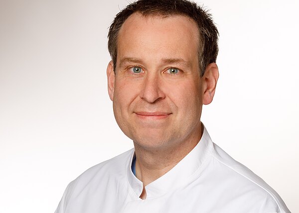 Porträt eines Mannes mittleren Alters mit kurzem braunem Haar. Er schaut lächelnd in die Kamera und trägt einen weißen Kittel, vor neutral hellem Hintergrund.