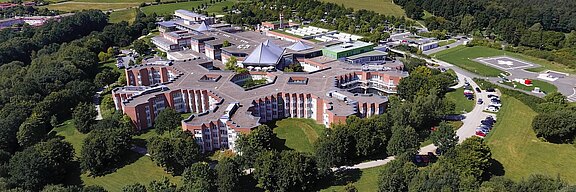 Luftaufnahme eines modernen Gebäudekomplexes, umgeben von Bäumen und Wiesen. Der Gebäudekomplex hat eine ungewöhnliche, sternförmige Architektur und beherbergt mehrere Flügel. Im Hintergrund sind weitere Einrichtungen und ein Landeplatz für Hubschrauber zu sehen.