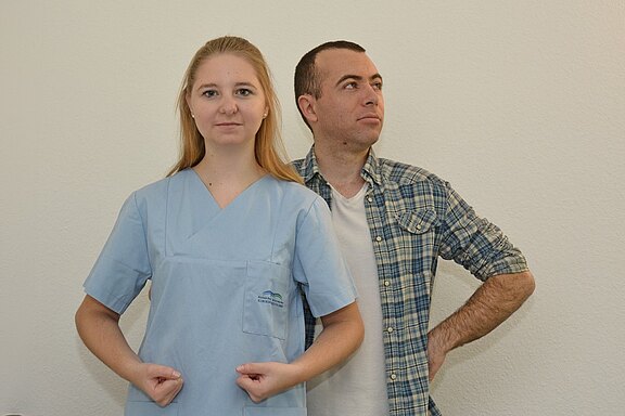 Eine Frau in einem blauen Pflegeanzug steht mit entschlossenem Gesichtsausdruck und geballten Fäusten auf der linken Seite, während ein Mann mit seriösem Blick und verschränkten Händen sich hinter ihr steht. Der Hintergrund ist hell und einfarbig.