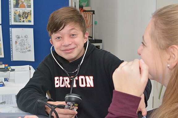 Ein lächelnder Junge mit einem Stethoskop um den Hals sitzt an einem Tisch. Ihm gegenüber sitzt ein Mädchen, das ihn anlächelt. Auf dem Tisch sind medizinische Utensilien und Unterlagen zu sehen. Im Hintergrund sind einige Bilder und Dokumente an einer Wand befestigt.