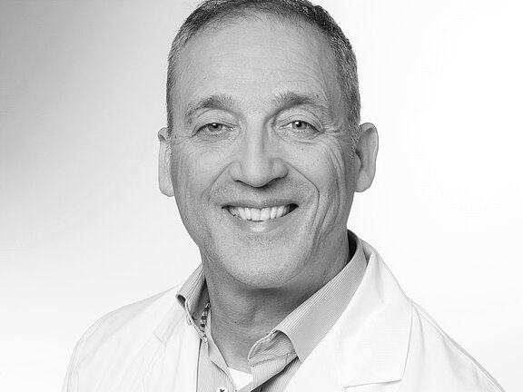 Schwarzweiß-Porträt: Ein lächelnder Mann mittleren Alters trägt einen weißen Arztkittel und Hemd. Neutrales, heller Hintergrund; Kopf und Oberkörper im Bild. Professioneller, freundlicher Ausdruck.