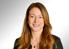 Porträtfoto einer lächelnden Frau mit langen, hellbraunen Haaren. Sie trägt ein dunkles Oberteil, eine Halskette und kleine Creolen. Heller, neutraler Hintergrund und ein freundlicher, professioneller Ausdruck.