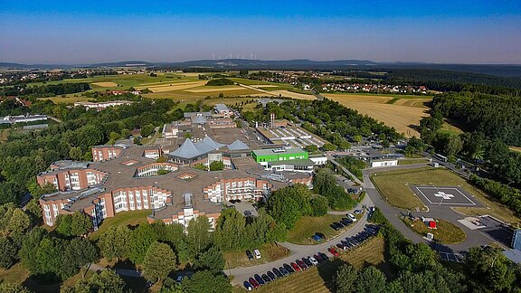 Luftaufnahme eines großen Krankenhauskomplexes mit mehreren Gebäuden, Parkplätzen und einem Helipad; umgeben von Feldern, Wald und einer kleinen Ortschaft am Horizont.