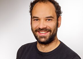 Porträt eines lächelnden erwachsenen Mannes mit dunklem, leicht lockigem Haar und Bart. Er trägt ein dunkles Shirt und schaut direkt in die Kamera, vor neutralem Hintergrund.