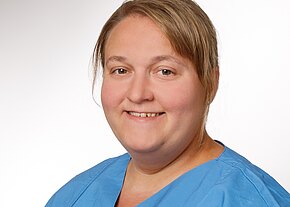 Kopf-und-Schulter-Porträt einer lächelnden Person in blauer medizinischer Kleidung vor neutralem Hintergrund.