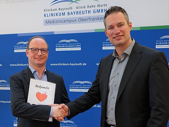 Zwei Männer beim formellen Händedruck; einer hält eine Urkunde mit Herzsymbol. Im Hintergrund ein Klinikbanner des Klinikums Bayreuth.