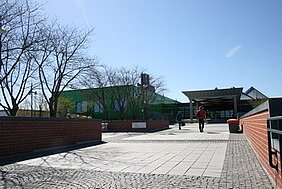 Das Bild zeigt einen gepflasterten Weg vor einem modernen Gebäude mit einem grünen Fassadenbereich. Menschen gehen entlang des Weges, bei klarem Himmel und Sonnenschein. Einige Bäume sind im Hintergrund sichtbar.