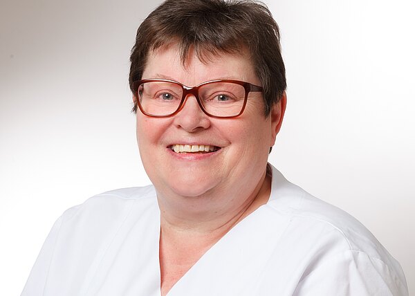 Porträt einer lächelnden Frau mit großer Brille, kurzen Haaren und weißer Berufsbekleidung vor hellem, neutralem Hintergrund; sie wirkt freundlich und professionell, wie eine Gesundheitsfachkraft.