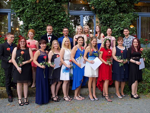 Gruppenfoto junger Erwachsener in festlicher Kleidung. Sie stehen in zwei Reihen im Freien, lächeln und halten rote Rosen sowie einige Urkunden. Der Anlass wirkt wie Abschlussfeier oder Ball.