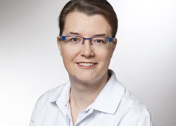 Porträt einer lächelnden erwachsenen Person mit zusammengebundenen Haaren und blauem Brillengestell. Sie trägt weiße, kittelähnliche Kleidung vor neutralem Hintergrund und blickt direkt in die Kamera.