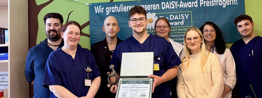 Gruppenfoto in einer Klinik: Ein Pflegeteam posiert, eine Person in der Mitte hält Urkunde und Trophäe. Dahinter hängt ein Banner zum DAISY-Award mit Glückwunschtext. Die Szene zeigt eine Auszeichnung und gemeinsamen Stolz.