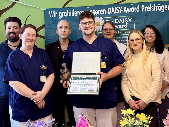 Gruppenfoto in einer Klinik: Ein Pflegeteam posiert, eine Person in der Mitte hält Urkunde und Trophäe. Dahinter hängt ein Banner zum DAISY-Award mit Glückwunschtext. Die Szene zeigt eine Auszeichnung und gemeinsamen Stolz.