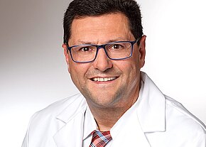 Porträt eines lächelnden Mannes mit kurzem dunkelbraunem Haar und blauer Brille, der einen weißen Arztkittel und eine Krawatte trägt; neutraler Hintergrund.