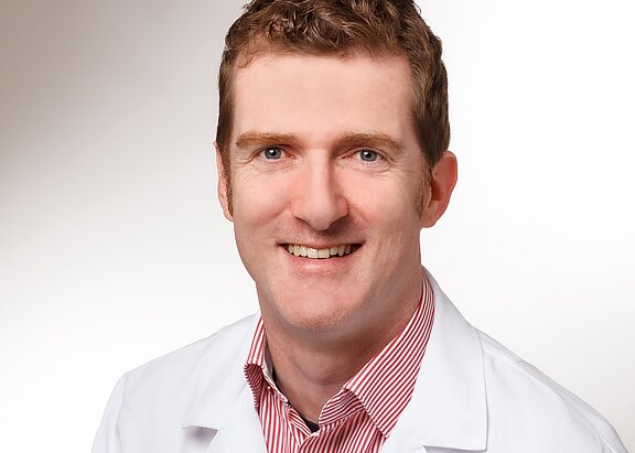 Porträt eines lächelnden Erwachsenen mit kurzem braunem Haar in weißem Laborkittel und gestreiftem Hemd vor neutralem Hintergrund. Kopf und Schultern sichtbar, freundlicher Blick direkt in die Kamera.