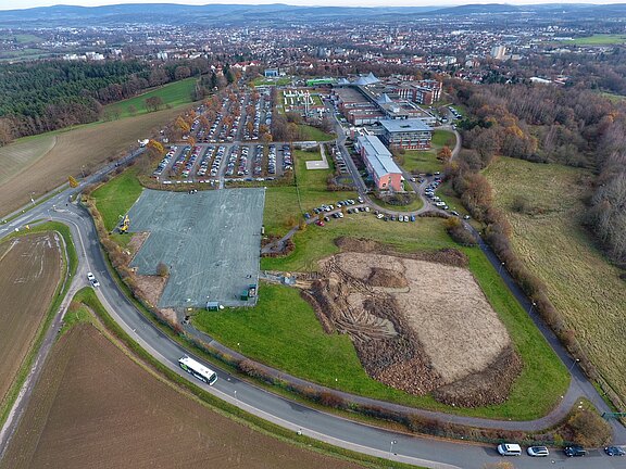 Luftaufnahme eines Klinik- oder Campusgeländes mit großen Parkplätzen, länglichen Gebäuden, angrenzenden Feldern und einer Baustelle im Vordergrund. Geschwungene Straßen umgeben das Areal; dahinter dichte Wohnbebauung und Hügel.