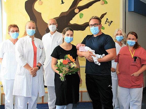 Eine junge Familie steht in einem Krankenhausflur. Die Mutter hält einen bunten Blumenstrauß, während der Vater ein neugeborenes Baby in den Armen trägt. Sechs Personen in weißen Kitteln und Masken stehen hinter ihnen, die offensichtlich medizinisches Personal sind.