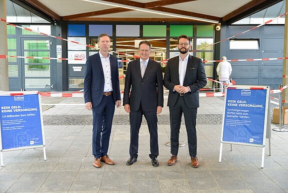 Drei Männer in Anzügen stehen vor einem mit Absperrband gesicherten Eingangsbereich. Links und rechts stehen blaue Schilder mit der Botschaft Kein Geld, keine Versorgung und Hinweisen auf fehlende Milliarden. Thema: Finanzierungslücken und Versorgungssicherheit.