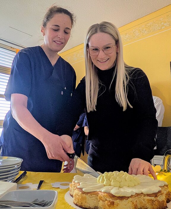 Zwei Personen schneiden gemeinsam eine Sahnetorte an; eine trägt dunkle Arbeitskleidung, die andere Brille. Auf dem Tisch liegen Teller. Fröhliche, helle Raumstimmung, Fokus auf gemeinsames Anschneiden des Kuchens.
